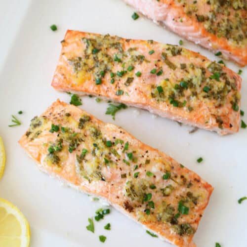 Three oven-baked salmon fillets topped with garlic and fresh herbs are arranged on a white plate, garnished with chives. Two lemon slices rest beside the salmon, enhancing this Oven-Baked Salmon with Lemon, Garlic, and Fresh Herbs dish.