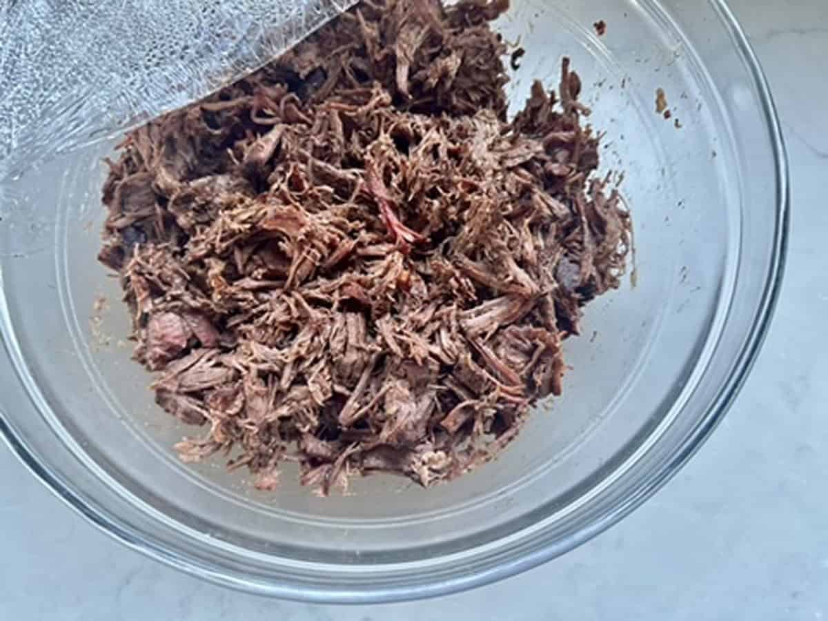 A clear glass bowl filled with shredded cooked meat sits on a light-colored surface, partially covered with plastic wrap.
