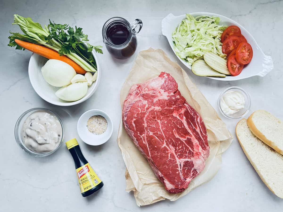 A slab of raw beef on parchment, surrounded by sliced onions, carrots, celery, lettuce, tomato, pickles, bread, mayonnaise, soup mix, seasoning, soy sauce, and a glass of dark liquid on a light countertop.