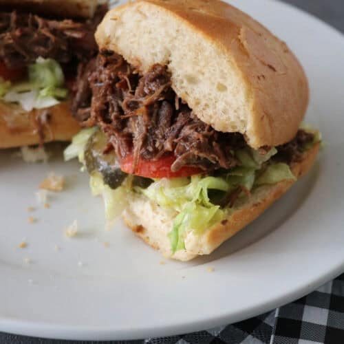 A shredded beef sandwich with lettuce, tomato, and pickles on a white plate, with a black and white checkered napkin beside it. The sandwich is cut in half, showing the filling.