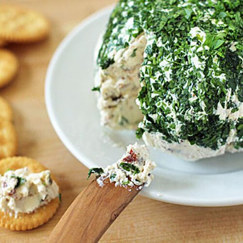 A wooden spreader lifts herbed cheese spread from a large cheese ball coated in chopped parsley, with round crackers arranged nearby on a wooden surface.