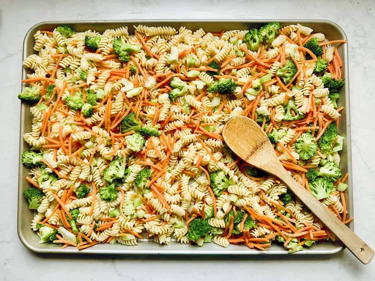A baking sheet filled with uncooked rotini pasta, broccoli florets, and julienned carrotsâperfect for a Pasta Salad with Broccoliârests under a wooden spoon.