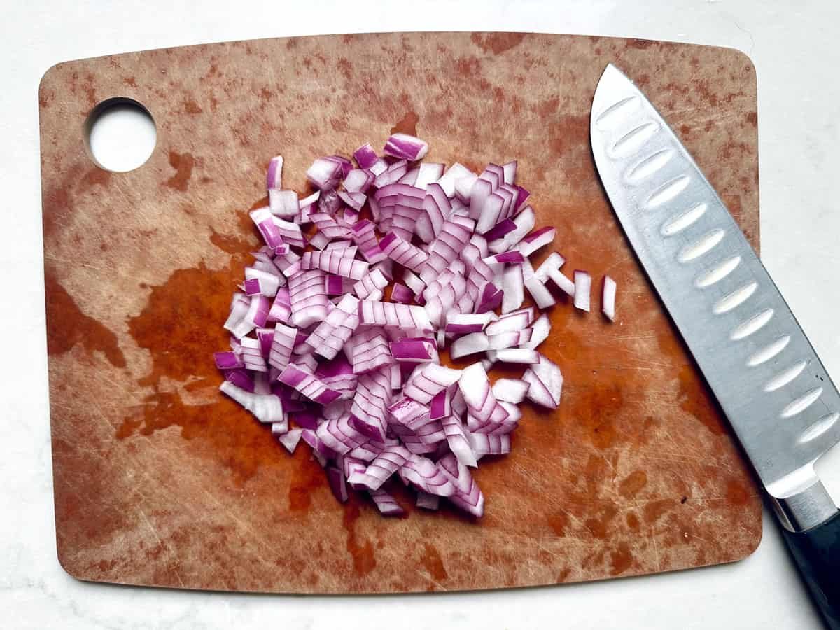 Finely diced red onion on a wooden cutting board with a chefâs knife beside it; the board shows juice stainsâperfect prep for Pasta Salad with Broccoli, Carrots, and PepperJack Cheese.