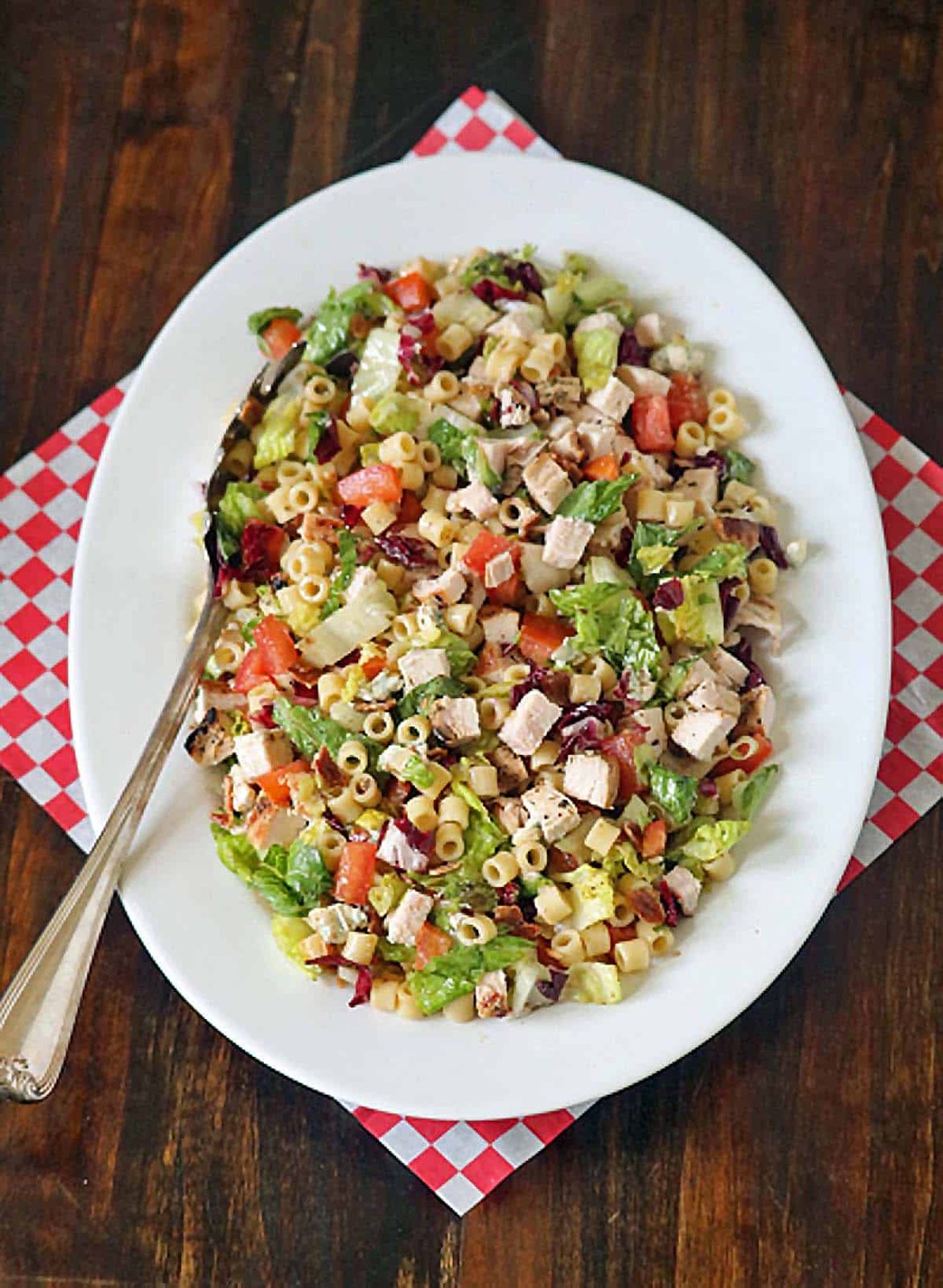 A white oval plate on a wooden table holds a Portillo's Chopped Salad with pasta, lettuce, tomatoes, grilled chicken, and other vegetables. A serving spoon rests on the plate, and a red-and-white checkered napkin is underneath.