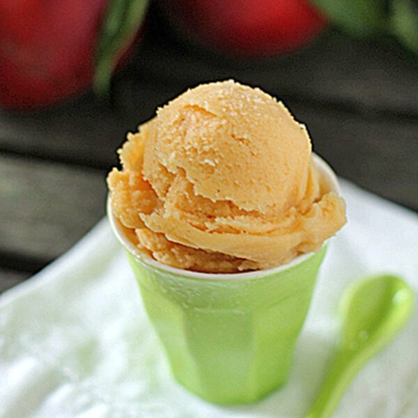 A scoop of peach sorbet sits in a light green cup on a white napkin, with a matching green spoon beside it. Out-of-focus peaches and green leaves are visible in the background.