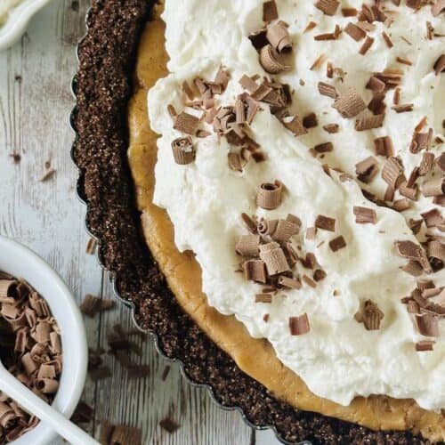 A close-up of a peanut butter chocolate tart with a chocolate crust, topped with whipped cream and chocolate shavings, sits on a rustic wooden surface. A small bowl filled with more chocolate shavings is nearby.