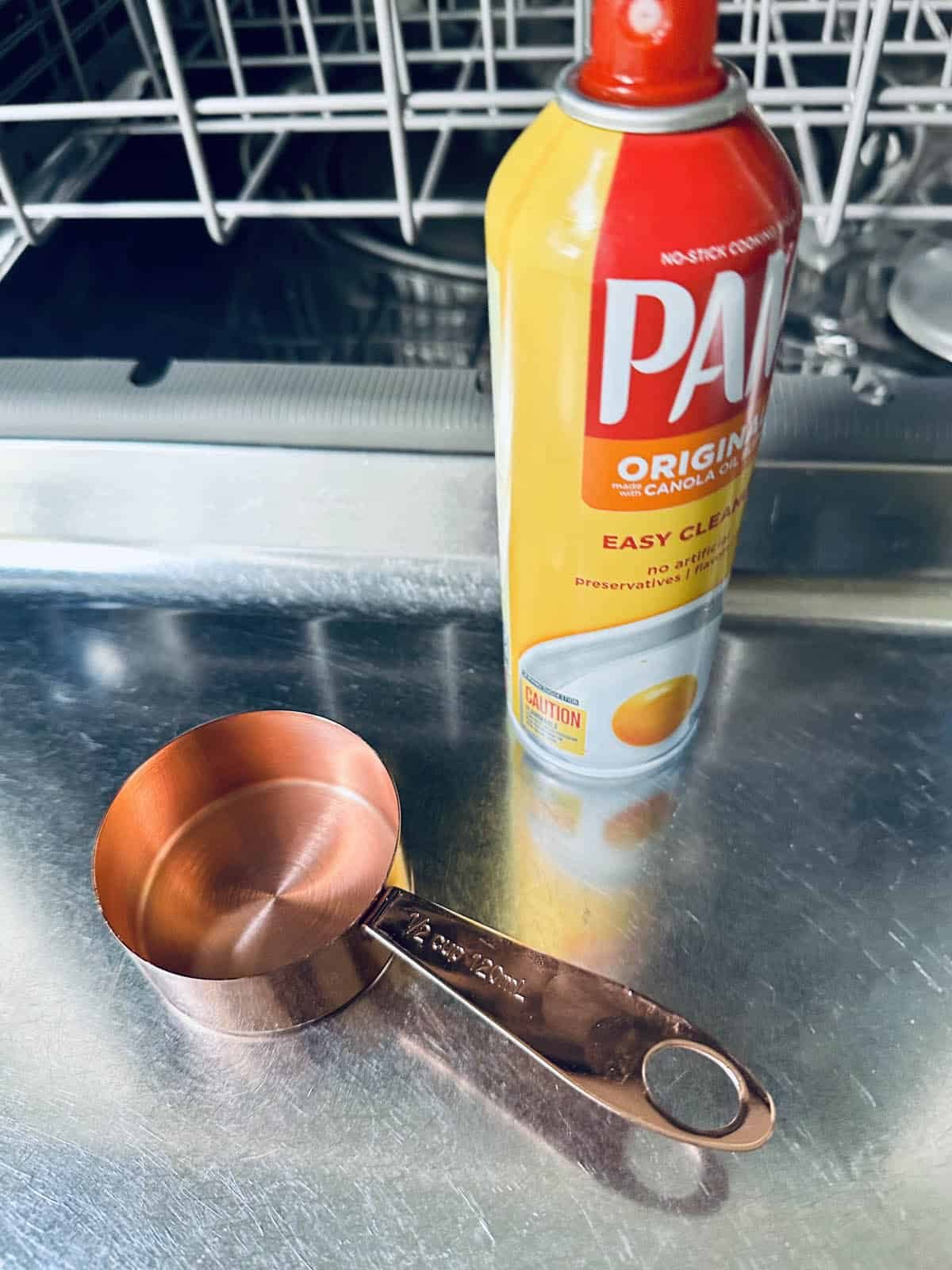 A can of PAM cooking spray and a copper measuring cup, ready for making peanut butter chocolate tart, sit on the open door of a dishwasher. The dishwasher's rack is visible in the background.