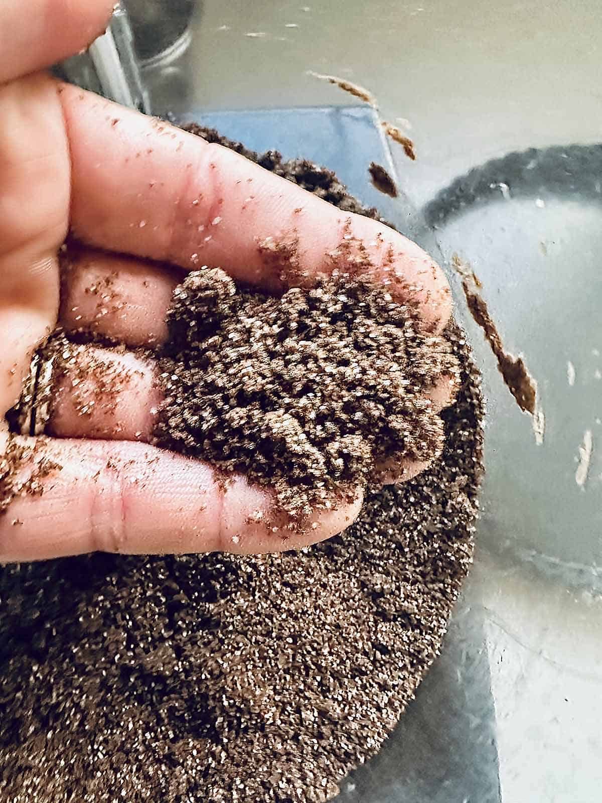 A close-up of a hand holding a small clump of dark, moist soil or sand—reminiscent of the rich layers in a peanut butter chocolate tart—with more of the same material below and a glass bowl in the background.