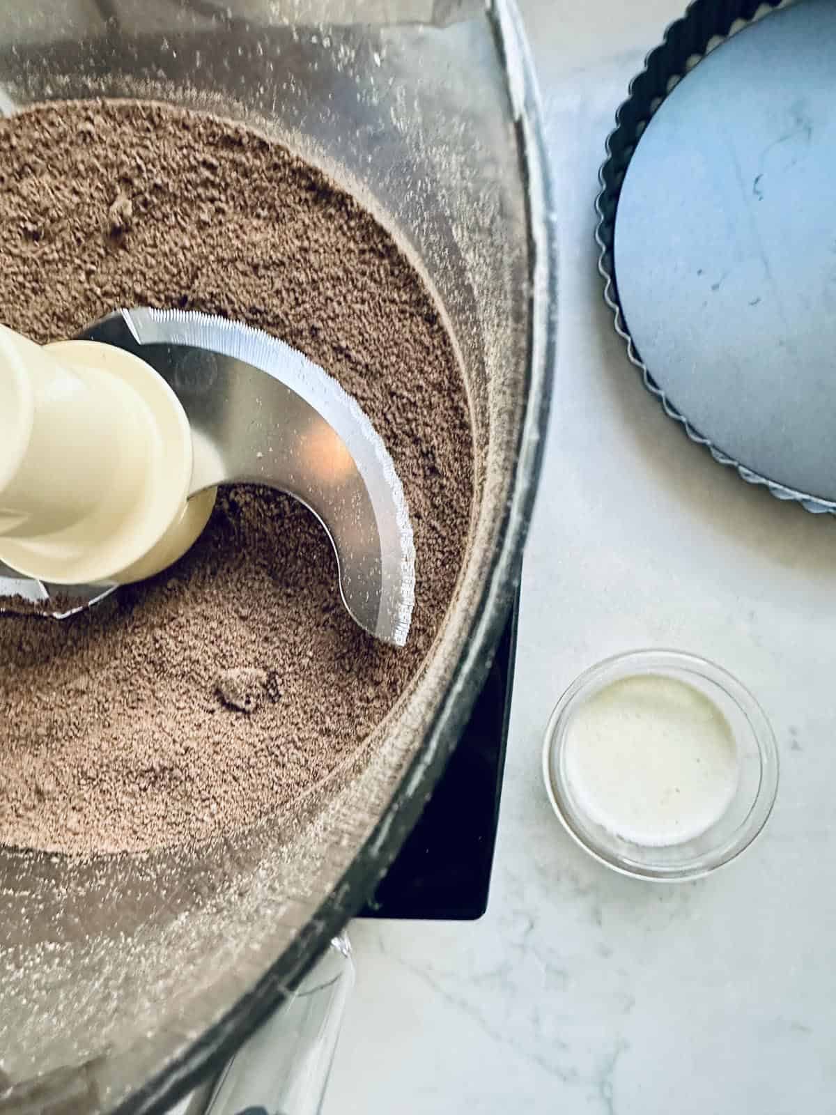 A food processor bowl with chocolate crumb mixture, a small glass bowl of liquid, and an empty tart pan on a light surface—perfect for making a peanut butter chocolate tart.