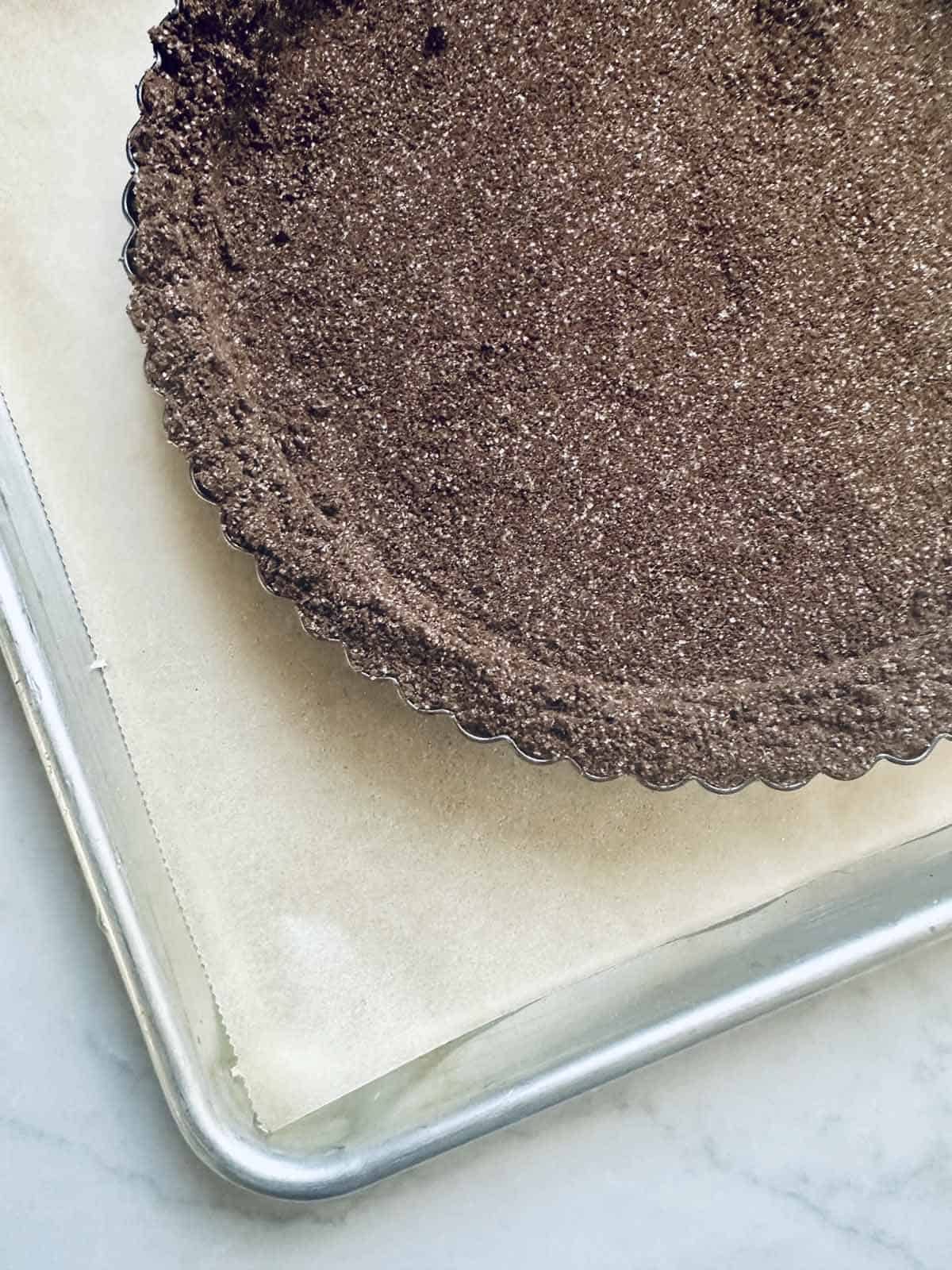 A close-up of a chocolate cookie crumb pie crust in a fluted tart pan, perfect as the base for a decadent peanut butter chocolate tart, resting on a parchment-lined metal baking sheet on a light-colored surface.