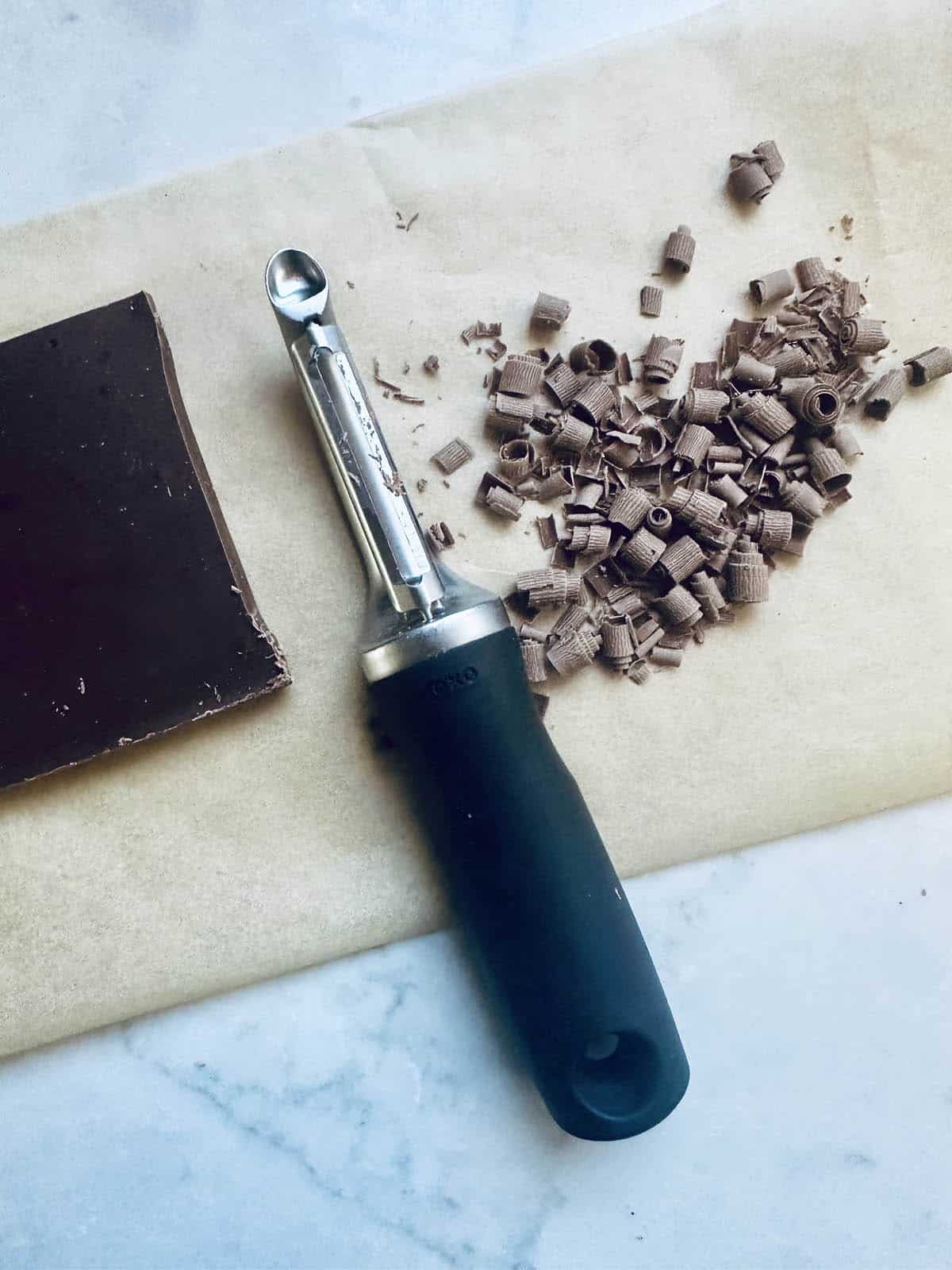 A vegetable peeler rests on parchment paper next to a block of chocolate and a small pile of chocolate curls—perfect for garnishing a decadent peanut butter chocolate tart.