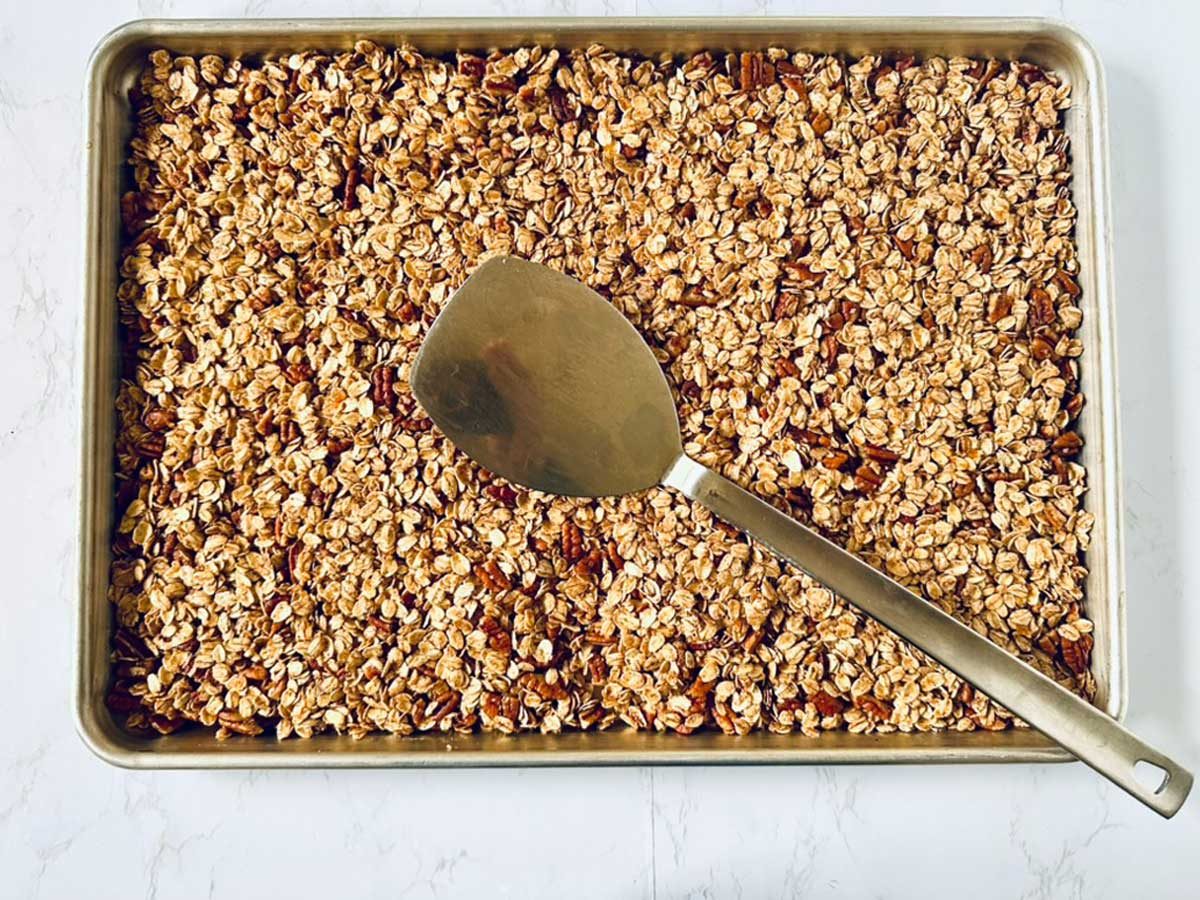 A baking sheet filled with toasted oats and nuts, likely granola, sits on a white surface. A large metal spatula rests on top of the granola.