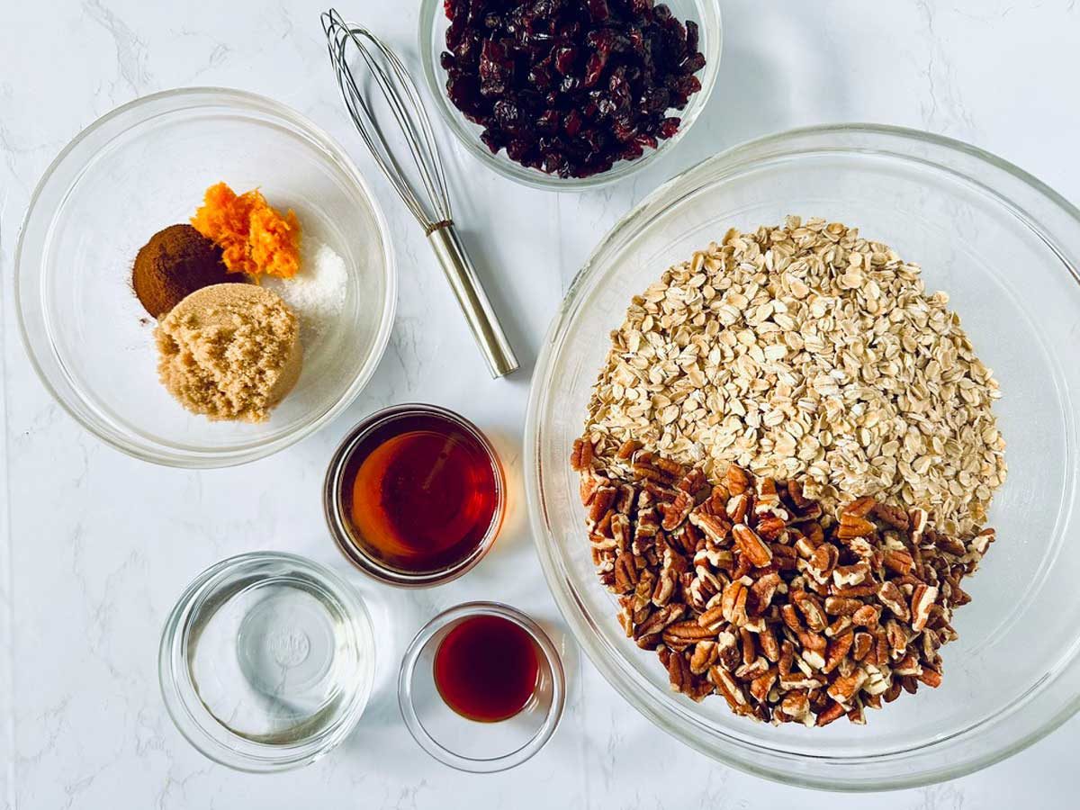 A top view of bowls containing oats, chopped pecans, dried cranberries, brown sugar, spices, orange zest, maple syrup, water, and vanilla extract, with a small whisk on a white marble surface.