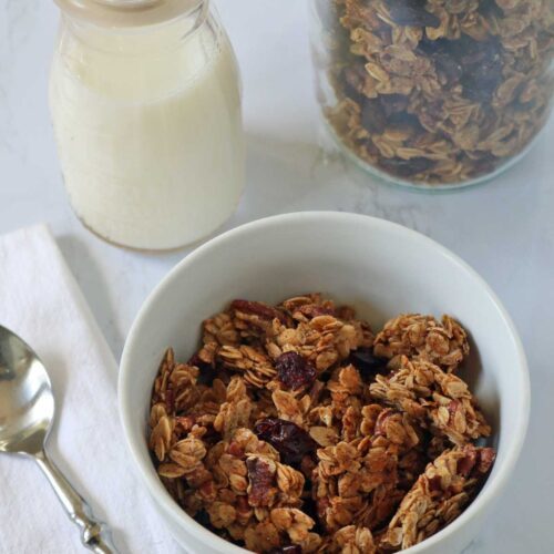 A white bowl of granola sits next to a glass jar of milk, a metal spoon, and a large jar of granola, all placed on a white surface with a folded white napkin.