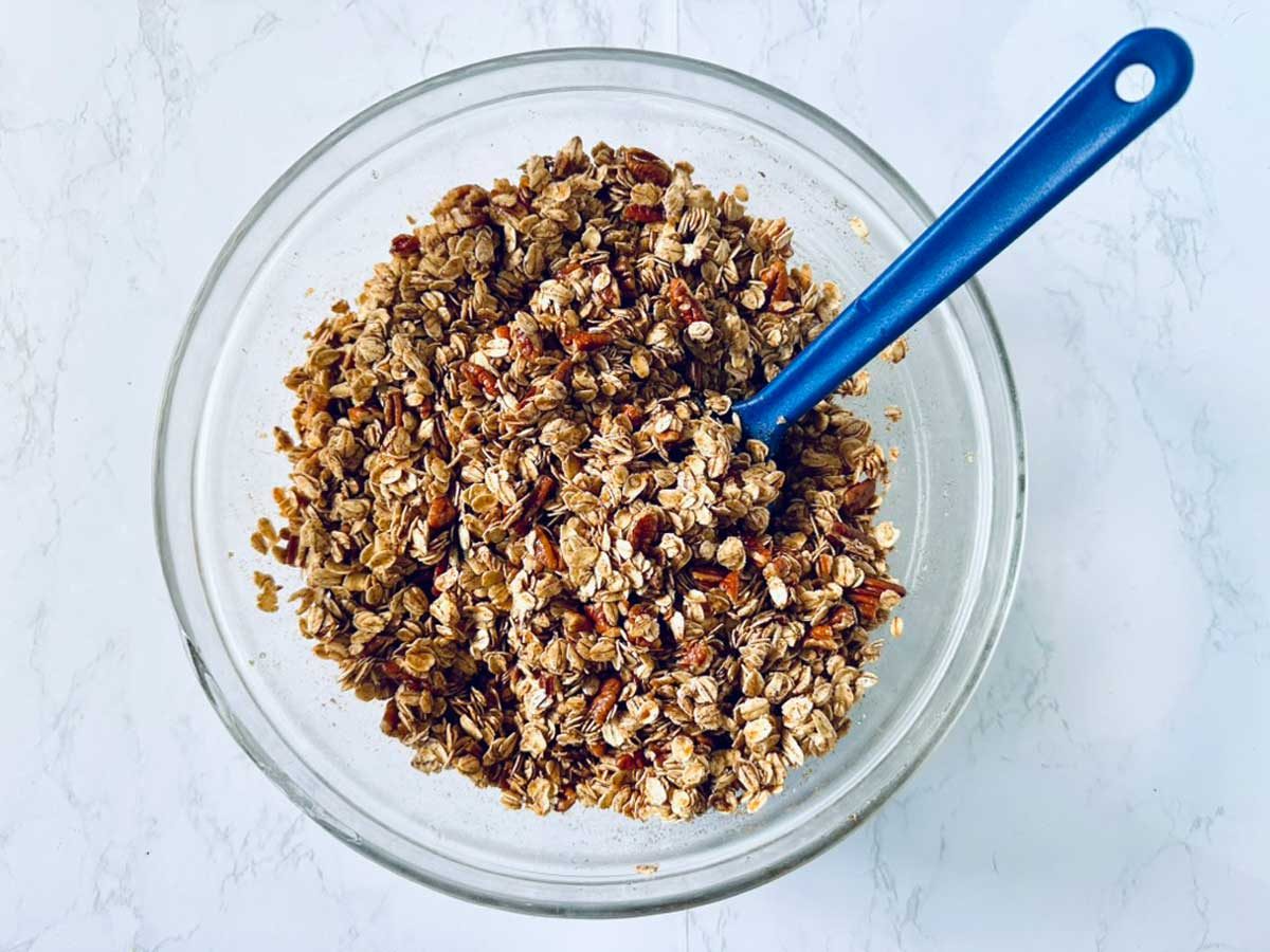 A glass bowl filled with granola mixture containing oats and nuts, with a blue spoon resting inside, on a white marble surface.