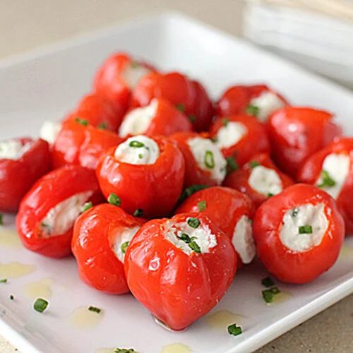A white plate with bright red cherry peppers stuffed with creamy white cheese, garnished with chopped green herbs and a drizzle of olive oil. The peppers are arranged in a pile, ready to be served as appetizers.