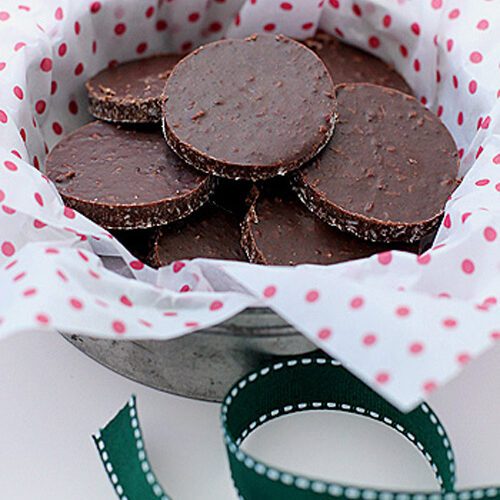 A metal bowl lined with white and pink polka dot paper holds round, chocolate cookies. A green ribbon with white stitching lies in front of the bowl.