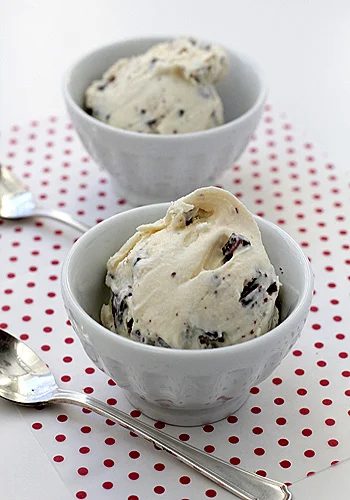 Two white bowls filled with vanilla ice cream with chocolate chunks are placed on a red and white polka dot surface. Two silver spoons lie beside the bowls.