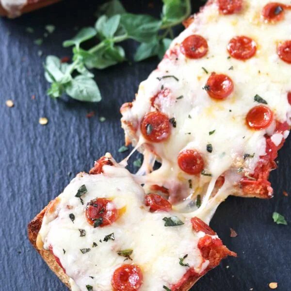 A slice of Pepperoni French Bread Pizza topped with melted cheese, mini pepperoni, and herbs is being pulled away, showing stretchy cheese. Fresh oregano leaves are scattered nearby on a dark slate surface.