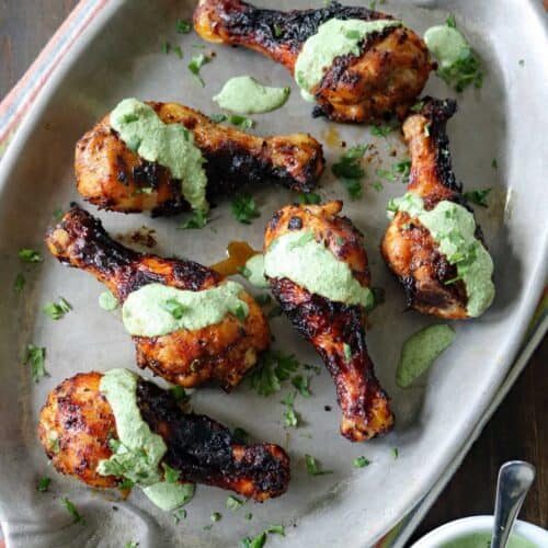 Air-Fried Peruvian Chicken with Green Crema drumsticks are arranged on a large oval platter, topped with vibrant green sauce and garnished with chopped herbs. A small bowl of extra green crema sits nearby on a striped cloth.