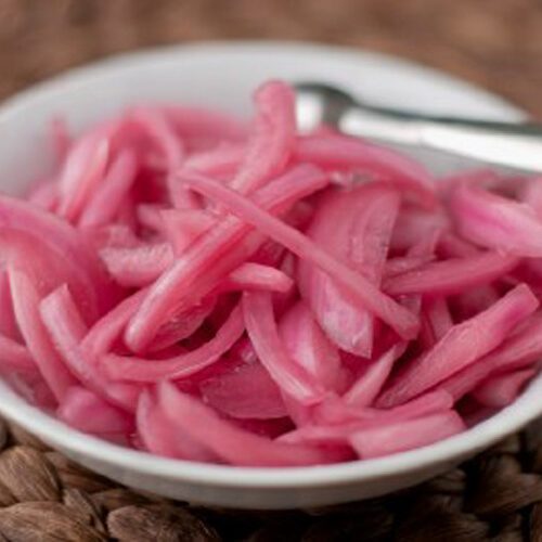 A white bowl filled with thinly sliced pickled red onions, with a metal fork resting on the bowl, placed on a woven brown mat.