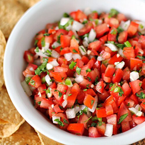 A white bowl filled with fresh pico de gallo made of diced tomatoes, onions, cilantro, and jalapeños, next to a pile of tortilla chips.