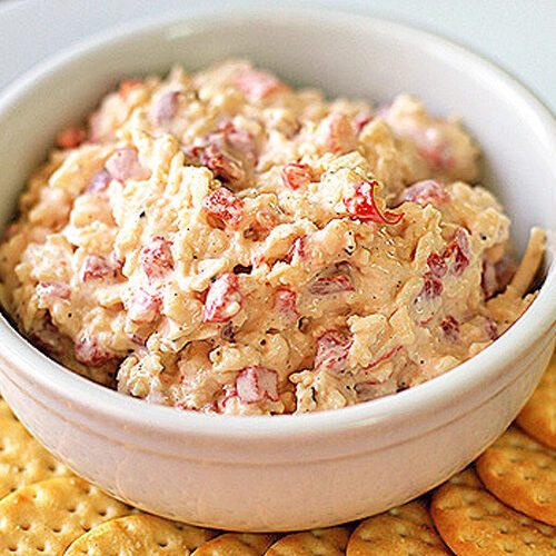 A white bowl filled with pimento cheese spread sits on a white plate, surrounded by round, golden crackers.