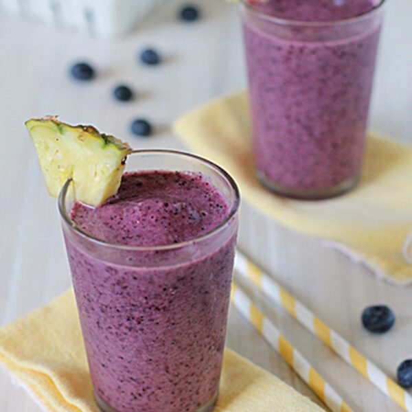 Two glasses of purple blueberry smoothie, each garnished with a pineapple wedge, sit on yellow napkins. Striped straws and scattered blueberries are nearby, with a container of blueberries in the background.