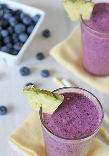 A glass of purple blueberry smoothie is garnished with a pineapple wedge and set on a yellow napkin. Another smoothie and a bowl of fresh blueberries are visible in the background.