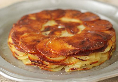A golden-brown, crispy potato galette sits on a silver plate, with thinly sliced potatoes layered in a circular pattern and lightly sprinkled with salt.