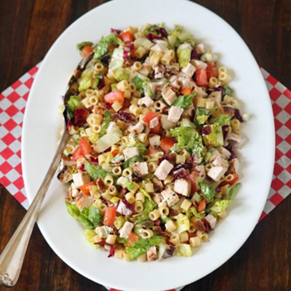 A white oval plate with chopped salad including pasta, lettuce, diced chicken, tomatoes, and vegetables, sits on a red and white checkered napkin with a serving spoon on a wooden table.