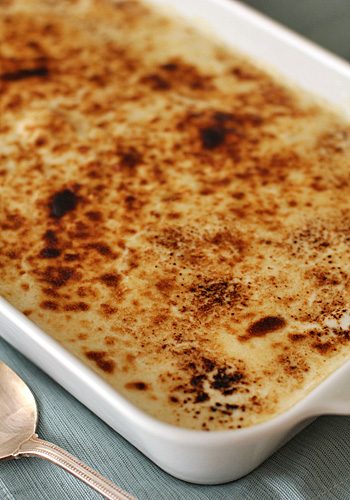 A close-up of a rectangular baking dish filled with a creamy dessert, topped with a caramelized, golden-brown crust. A silver spoon rests beside the dish on a light blue tablecloth.