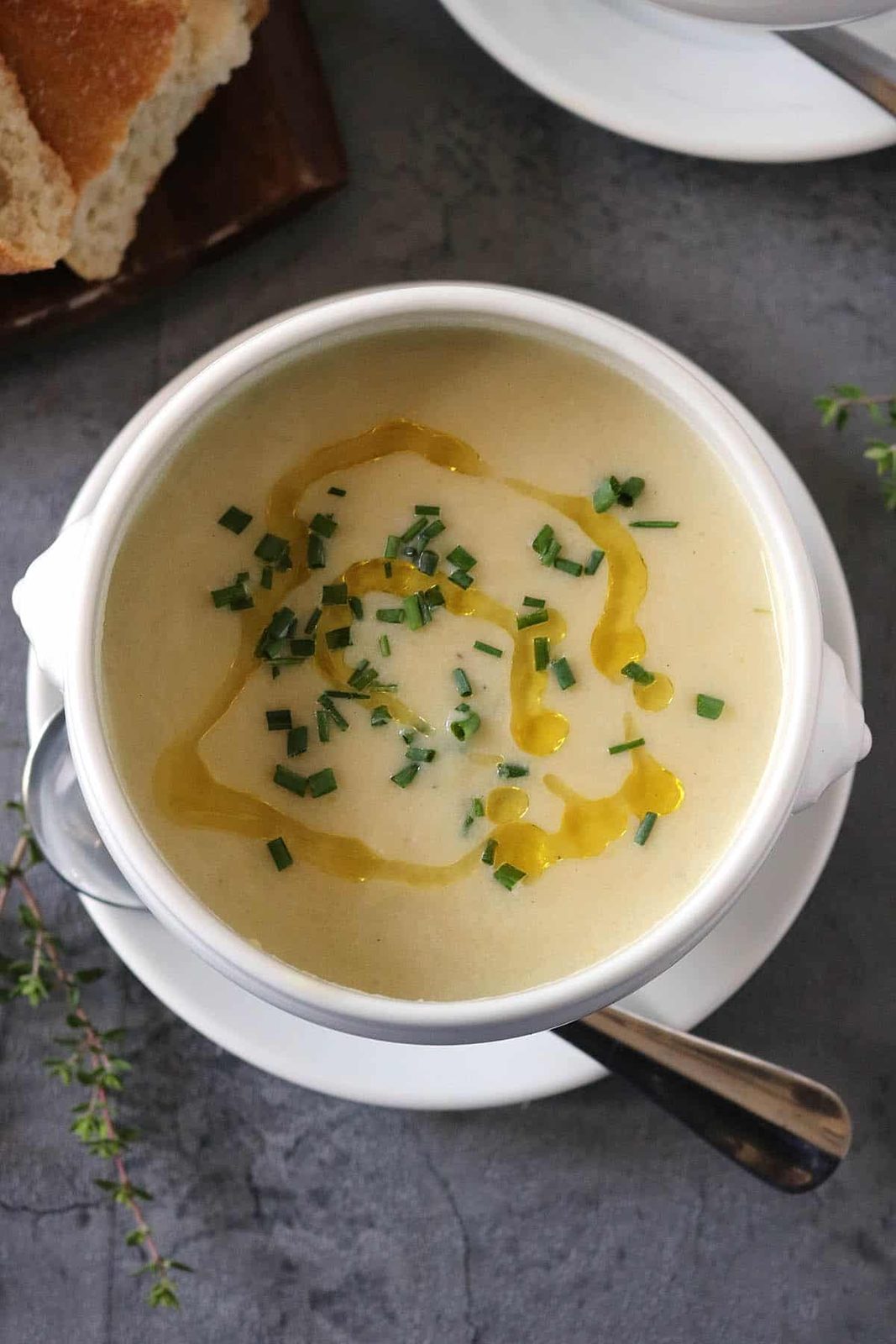 A bowl of creamy Potato Leek Soup garnished with chopped chives and a drizzle of olive oil, served with a spoon on a white saucer. Bread is visible in the background on a dark surface.