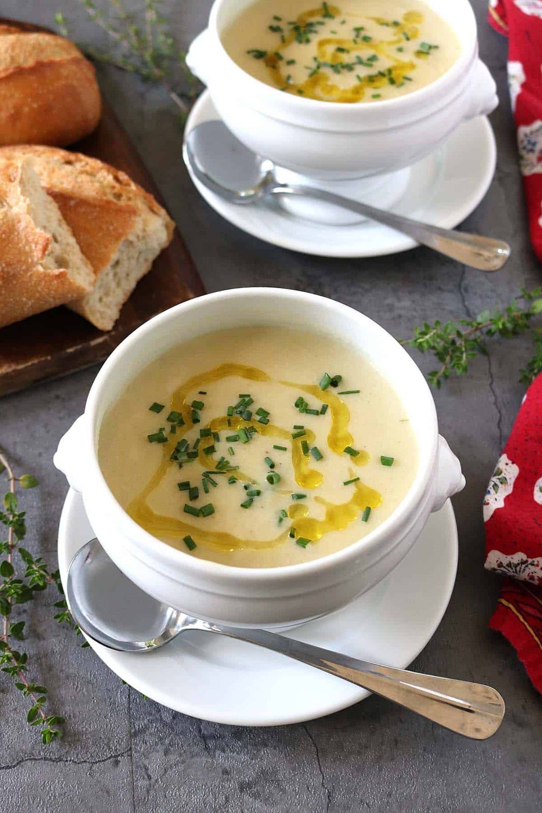 Two white bowls of creamy Potato Leek Soup topped with chopped herbs and a drizzle of oil, each on a saucer with a spoon. A loaf of crusty bread is on a wooden board nearby. Fresh herbs and a red napkin decorate the table.