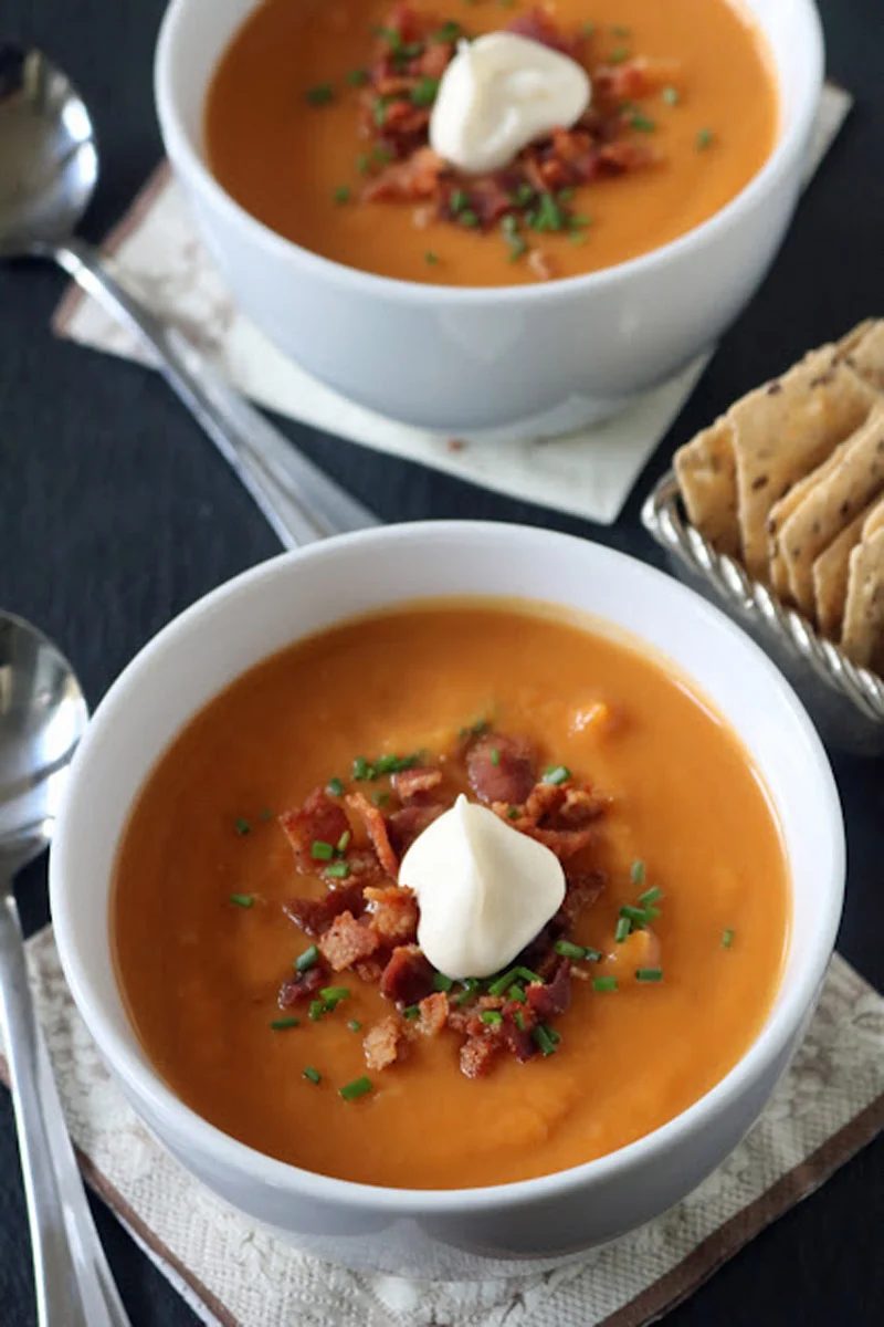 Two bowls of creamy orange soup topped with bacon bits, chopped chives, and a dollop of sour cream. Spoons, crackers, and napkins are placed nearby on the table.