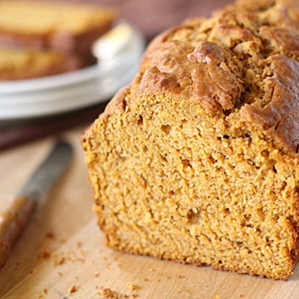 A close-up of a loaf of pumpkin bread on a wooden surface, with a knife beside it and a plate of sliced bread in the background. The bread has a golden-brown crust and moist, textured interior.