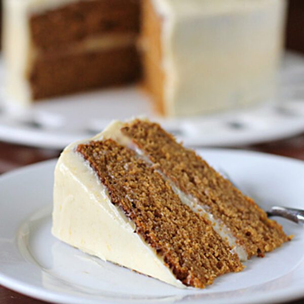 A slice of two-layer carrot cake with cream cheese frosting sits on a white plate with a fork. The rest of the cake is in the background on a cake stand.