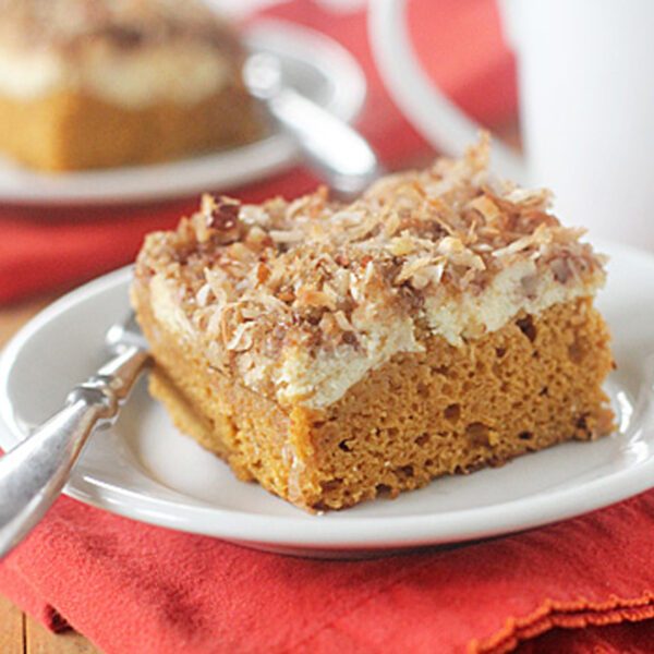 A slice of pumpkin cake topped with cream cheese and toasted coconut sits on a white plate with a silver fork, placed on a red napkin. Another plate with cake and a white mug are visible in the background.
