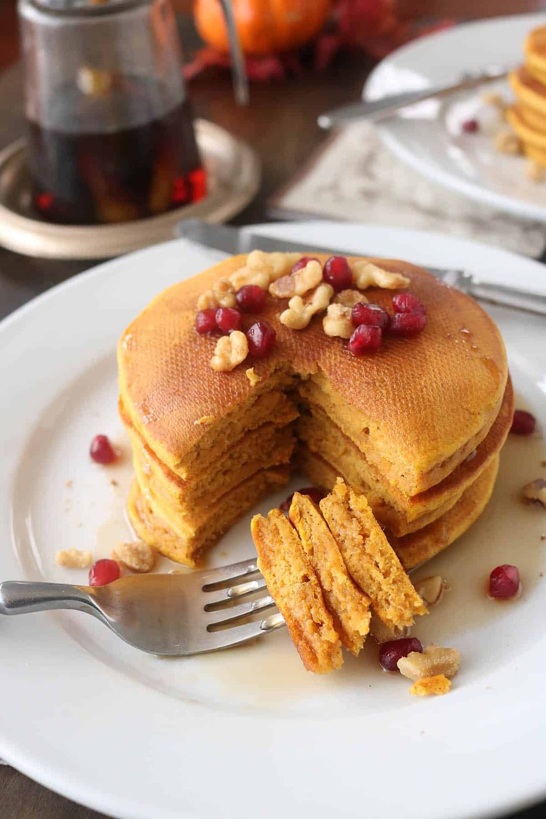 A stack of pumpkin pancakes topped with walnuts and pomegranate seeds sits on a white plate. A fork holds a cut bite, and syrup is drizzled over the pancakes. A syrup jar is in the background.