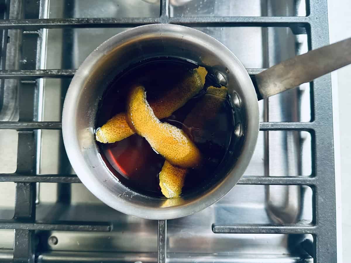 A metal pot with dark liquid and several orange peels sits on a stovetop burner, viewed from above.