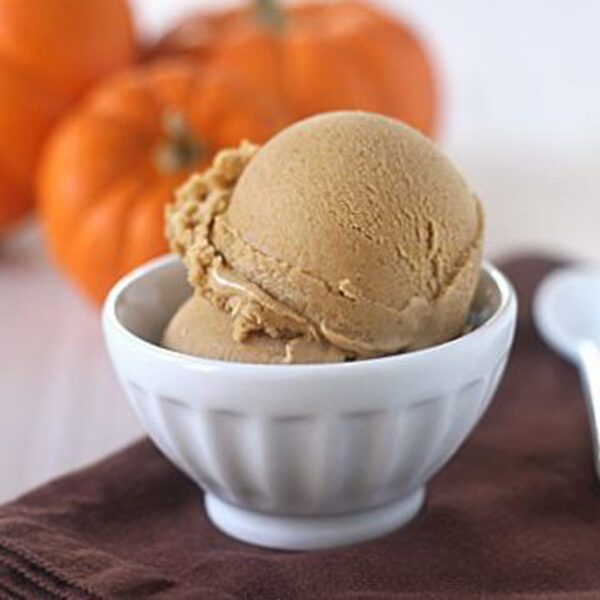 A white bowl holds two scoops of brown ice cream, placed on a folded brown cloth with a white spoon nearby. Three small orange pumpkins are in the blurred background.