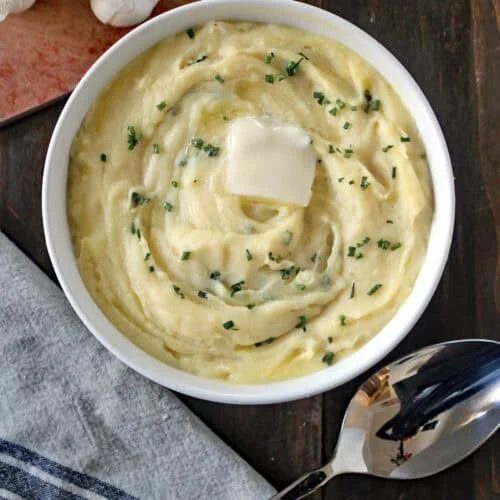 A bowl of creamy mashed potatoes topped with a pat of butter and chopped chives, placed on a wooden table next to a spoon, garlic, and a small dish of chopped herbs.