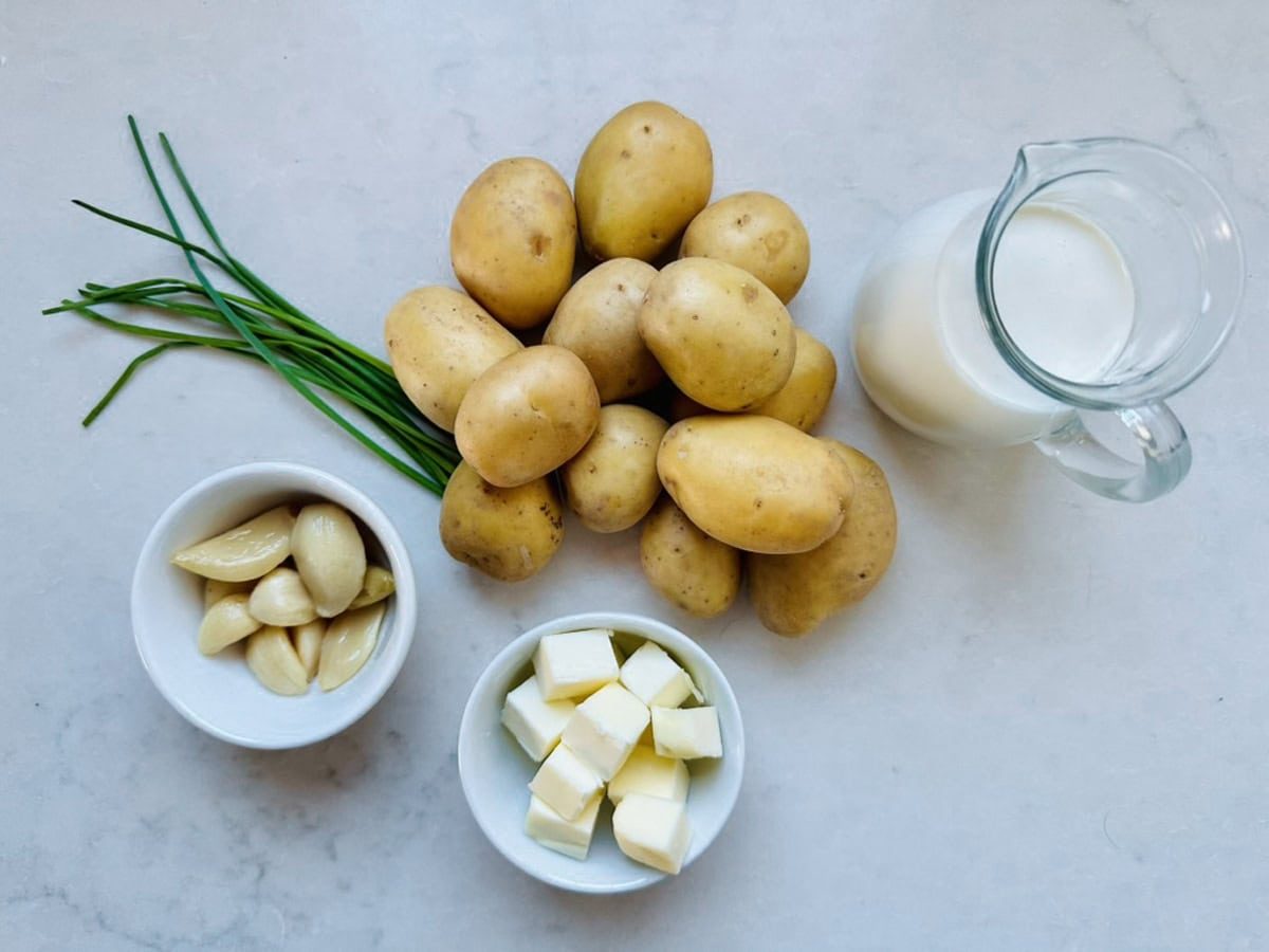 Several whole potatoes, a bunch of chives, a small bowl of peeled garlic cloves, a bowl of cubed butter, and a glass pitcher of milk arranged on a white surface.