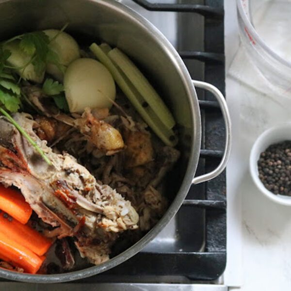 A pot on a stove filled with chicken bones, carrots, celery, onions, and parsley, ready to make broth. Nearby on the counter are bowls of black peppercorns and salt, and an empty measuring cup.
