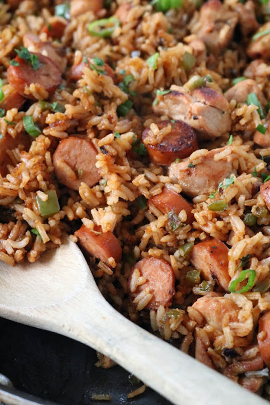 A close-up of a wooden spoon stirring a skillet filled with jambalaya, featuring cooked rice, sliced sausage, diced chicken, green onions, and herbs.