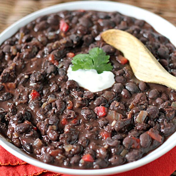 A bowl of black bean stew garnished with a dollop of sour cream and a sprig of cilantro, with a wooden spoon resting on the edge and a red napkin underneath.