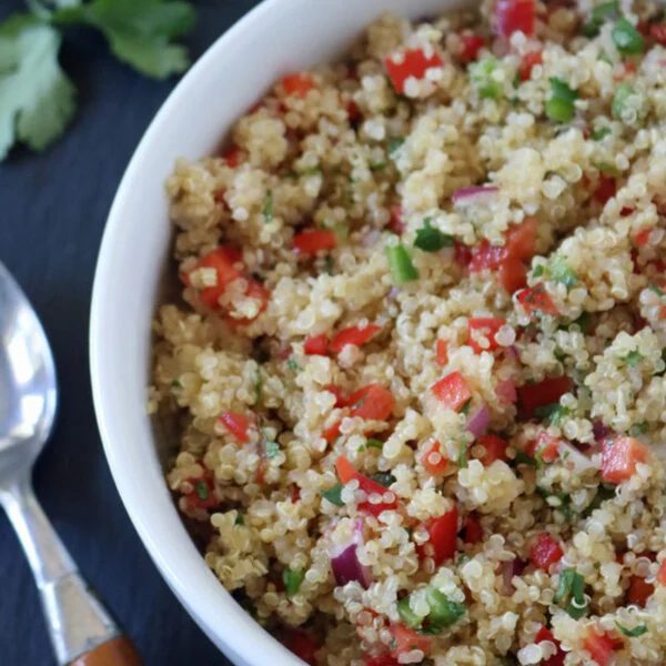 A white bowl filled with quinoa salad mixed with chopped red bell pepper, red onion, and green herbs sits next to a bamboo-handled spoon on a dark surface, with fresh cilantro leaves nearby.