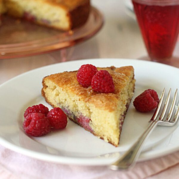 A slice of golden cake with raspberries on top and on the side, served on a white plate with a fork. A glass of raspberry drink and the rest of the cake are visible in the background.