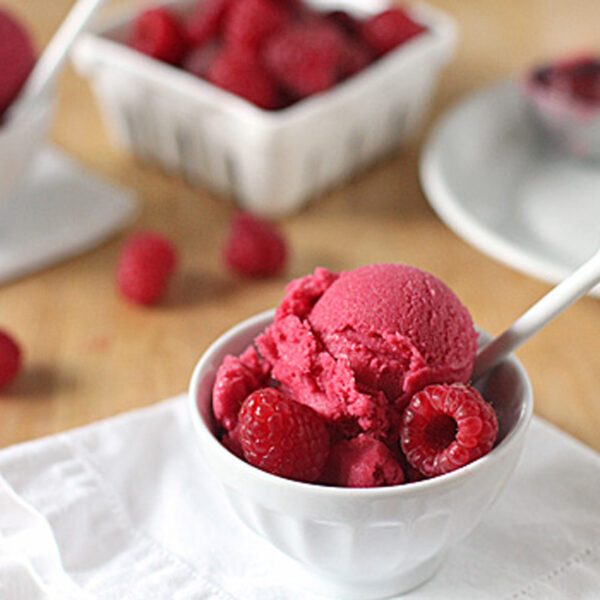 A white bowl filled with scoops of raspberry sorbet, garnished with fresh raspberries and a white spoon. In the background, there are more raspberries and a white container on a wooden table.