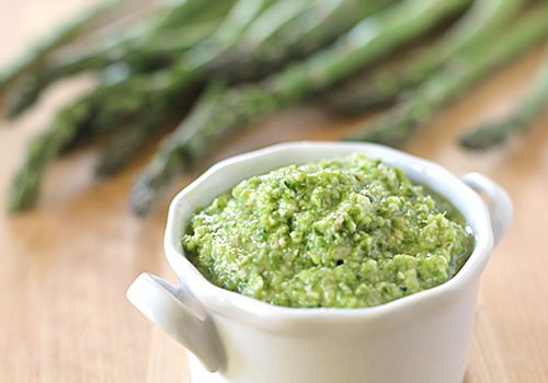 A small white bowl filled with green asparagus pesto sits on a wooden surface, with fresh asparagus spears blurred in the background.